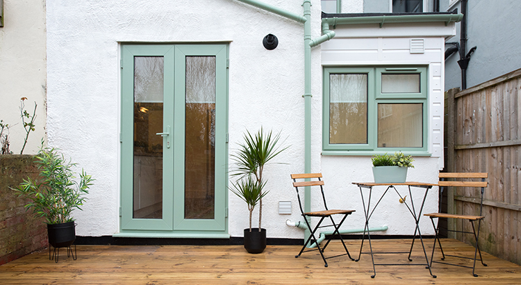 A general exterior view of a baack garden patio area with wood decking, potted plants, Dragon palm tree, metal table and two chairs pale pastel sage green patio doors, window and drainpipe